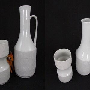 Könnte beinhalten: Zwei weiße Keramikvasen mit einem strukturierten Design. Eine Vase ist hoch und schlank mit einem Henkel, die andere ist kürzer und breiter mit einer zylindrischen Form.
