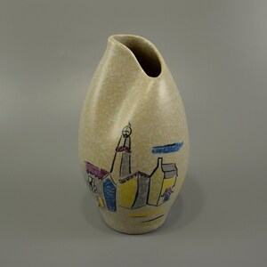 Könnte beinhalten: Eine beige Keramikvase mit einer einzigartigen, wellenförmigen Form. Sie zeigt ein handgemaltes Design einer kleinen Stadt mit bunten Gebäuden und einem Kirchturm.