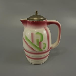 May include: A white ceramic pitcher with a pink and green geometric design. The pitcher has a metal lid and a handle.