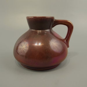 Könnte beinhalten: Ein brauner Keramik Krug mit rundem Körper und einem einzigen Henkel. Der Krug hat eine glänzende Oberfläche und einen dezenten irisierenden Schimmer.