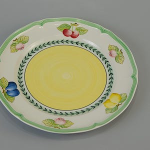 May include: A white ceramic plate with a scalloped edge and a yellow center. The plate is decorated with green trim and colorful fruit illustrations, including cherries, plums, and lemons. A decorative item for the kitchen or dining room.