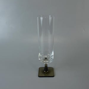 Könnte beinhalten: Ein hohes, klares Glas mit einem dunklen Stiel und einem quadratischen Sockel. Das Glas hat eine zylindrische Form und steht auf einer grauen Oberfläche. Der Sockel ist dunkel und quadratisch. Das Glas ist leer.