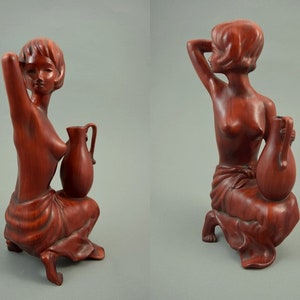 May include: A wooden sculpture of a woman sitting with a pitcher. The sculpture is painted in a reddish-brown color.