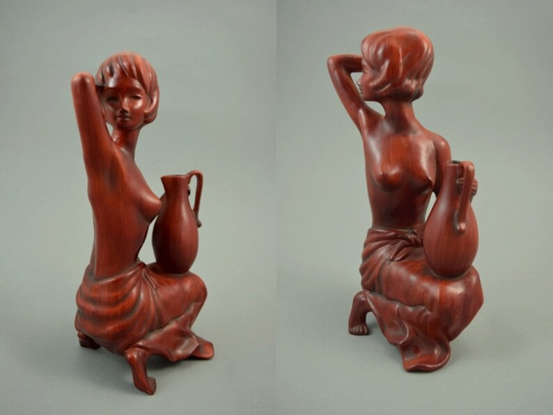 May include: A wooden sculpture of a woman sitting with a pitcher. The sculpture is painted in a reddish-brown color.