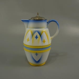 Pode incluir: Um jarro de cerâmica branca com um design geométrico azul e amarelo. O jarro tem uma tampa prateada e uma alça.