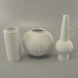 Può includere: Tre vasi in ceramica bianca con disegni in rilievo. I vasi sono di forme e dimensioni diverse. Il vaso a sinistra è cilindrico, il vaso al centro è rotondo e il vaso a destra è alto e sottile.