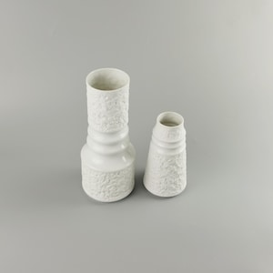 May include: Two white ceramic vases with an embossed floral design. The taller vase has a wider base and a narrower neck. The shorter vase has a more conical shape.