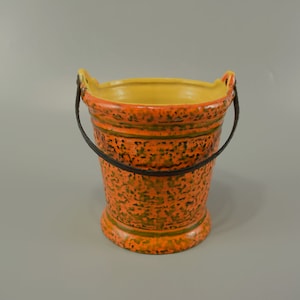 Könnte beinhalten: Orangener und gelber Keramik-Eimer mit schwarzen Griffen. Der Eimer hat eine strukturierte Oberfläche mit einem grün-schwarzen Muster.