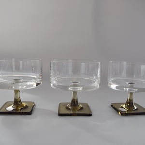 Könnte beinhalten: Drei klare Dessertschalen aus Glas mit quadratischem Fuß und kurzem Stiel. Die Schalen haben eine zylindrische Form und bestehen aus klarem Glas. Die Stiele und Basen sind olivgrün.