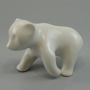 May include: A white ceramic figurine of a bear cub standing on all fours. The bear cub is looking down and has a rounded, smooth body.