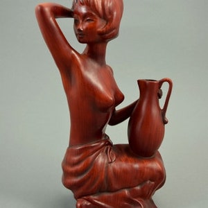 May include: A wooden sculpture of a woman holding a pitcher. The sculpture is made of a reddish-brown wood and is in a seated position. The woman is wearing a simple garment and has her hair pulled back.