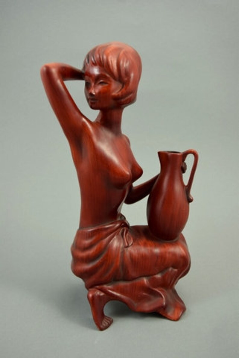 May include: A wooden sculpture of a woman holding a pitcher. The sculpture is made of a reddish-brown wood and is in a seated position. The woman is wearing a simple garment and has her hair pulled back.