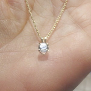 May include: A gold-colored necklace with a small, round, clear gemstone pendant. The necklace is held in a person's hand, showcasing the delicate chain and the sparkling pendant. The pendant is the focal point of the jewelry.
