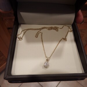 May include: A gold necklace with a small, clear gemstone pendant, displayed in a dark brown jewelry box with a cream-colored interior. The necklace chain is delicate and the pendant is oval-shaped.