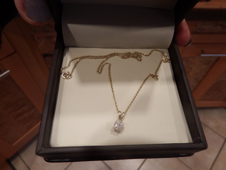 May include: A gold necklace with a small, clear gemstone pendant, displayed in a dark brown jewelry box with a cream-colored interior. The necklace chain is delicate and the pendant is oval-shaped.