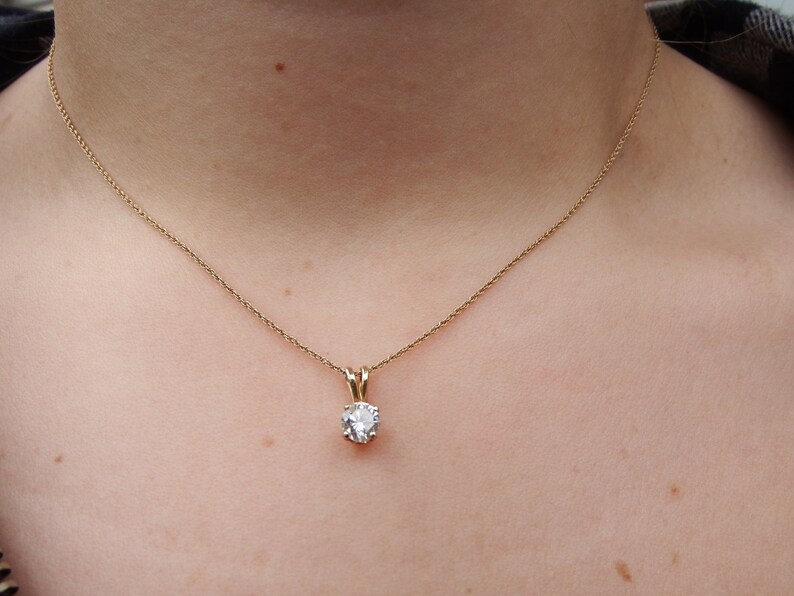 May include: A gold-colored necklace with a small, round, clear gemstone pendant. The pendant is held in place by a gold-colored setting. The necklace is displayed on a person's neck.