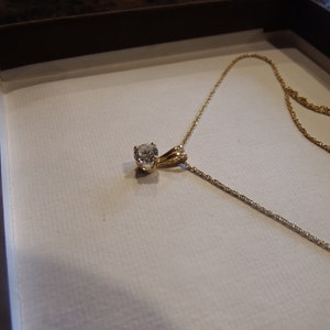 May include: A gold necklace with a round, clear gemstone pendant. The necklace is displayed in a white box with a brown border. The chain is delicate and appears to be made of gold.