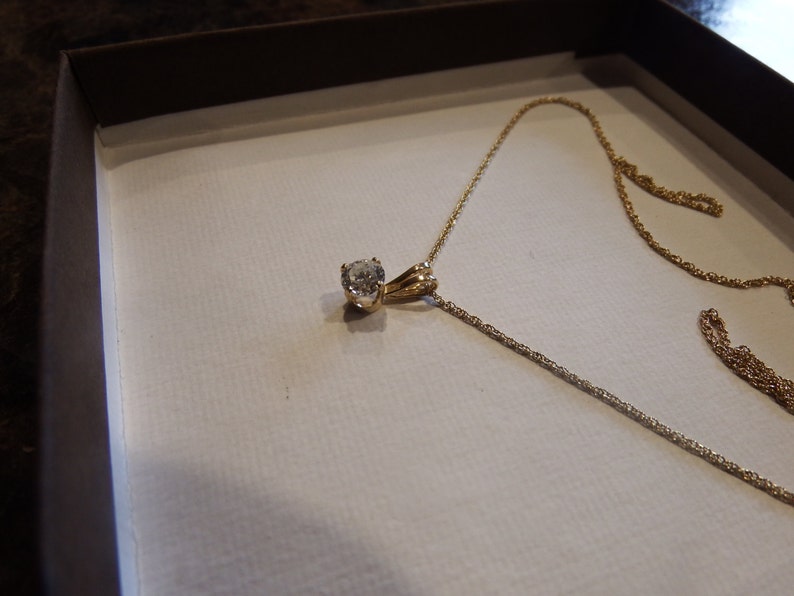 May include: A gold necklace with a round, clear gemstone pendant. The necklace is displayed in a white box with a brown border. The chain is delicate and appears to be made of gold.