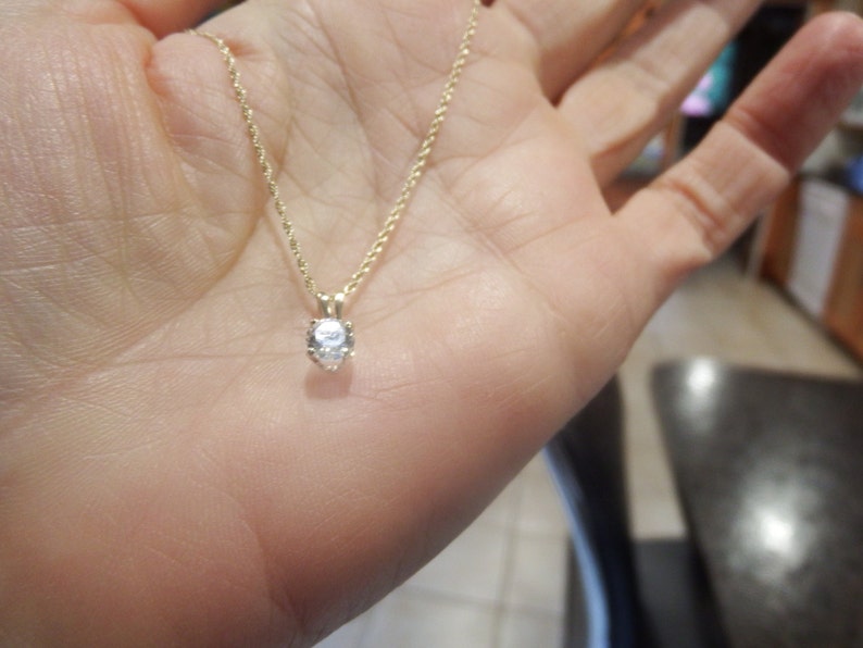 May include: A gold-colored necklace with a small, round, clear gemstone pendant. The necklace is held in a person's hand, showcasing the delicate chain and the sparkling pendant. The pendant is the focal point of the jewelry.