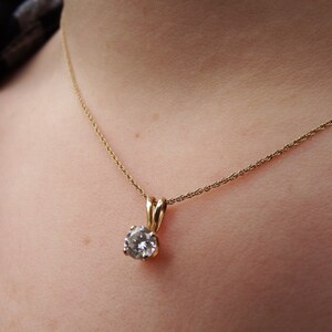 May include: A gold-colored necklace with a small, round, clear gemstone pendant. The pendant is held by a gold-colored setting. The necklace is worn around the neck.