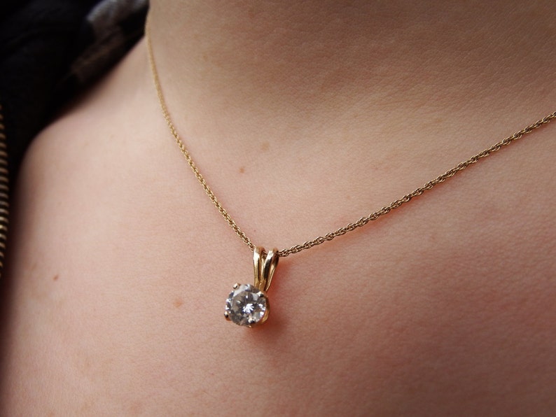 May include: A gold-colored necklace with a small, round, clear gemstone pendant. The pendant is held by a gold-colored setting. The necklace is worn around the neck.