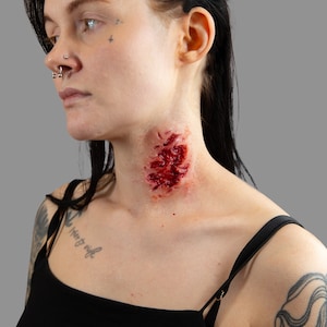 May include: A woman with black hair and tattoos has a large, bloody wound on her neck. The wound is red and raw, and there is blood dripping from it.