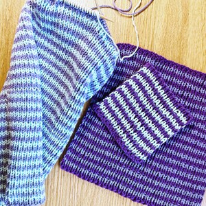 May include: Two pieces of knitted fabric in a striped pattern of purple and light blue. The fabric is laid out on a wooden surface.