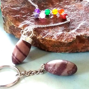Puede incluir: Un conjunto de colgante y llavero con piedras ovaladas a rayas en tonos marrón y crema. El colgante cuelga de una cadena plateada con cuentas de colores. El llavero tiene un anillo plateado. Los artículos se exhiben sobre una piedra marrón texturizada.