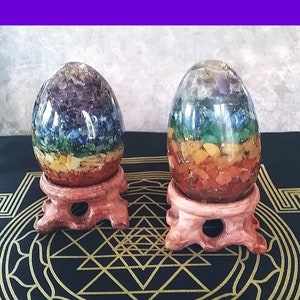 Puede incluir: Dos objetos decorativos en forma de huevo llenos de piedras preciosas de colores, dispuestas en bandas de color. Los huevos se asientan sobre pequeños soportes de madera marrón claro. El fondo es una tela verde oscuro con un patrón geométrico dorado, enmarcado por un borde púrpura.