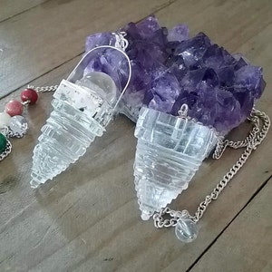 Puede incluir: Dos péndulos de cristal de cuarzo transparente con cadenas plateadas. Los péndulos tienen forma de pirámide con una punta puntiaguda. Los péndulos cuelgan de un racimo de cristales de amatista.