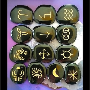 May include: Set of 12 black obsidian stones with gold symbols, including an arrow, a crescent moon, a sun, and a spiral.