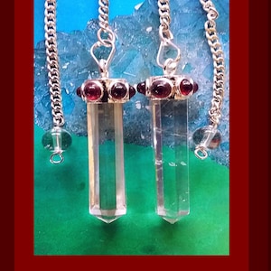May include: Two clear quartz crystal pendulums with silver chains and red gemstone accents. The pendulums are suspended against a blue and green background, framed by a red border. The crystals are faceted and elongated.