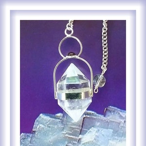 May include: A clear quartz crystal pendulum with a silver chain and a silver ring at the top. The pendulum is hanging over a blue and white crystal.