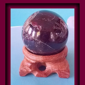Könnte beinhalten: Ein dunkelroter, polierter, kugelförmiger Edelstein auf einem Holzständer. Der Edelstein hat ein marmoriertes Muster.