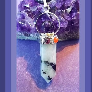 May include: A silver pendant with a clear crystal point, surrounded by a silver ring with a purple amethyst stone. The pendant is accented with red and orange stones and hangs from a black cord.