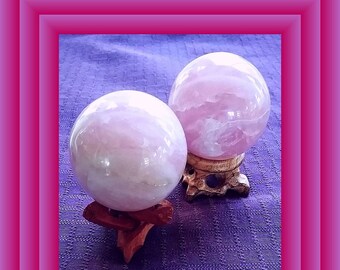 Könnte beinhalten: Zwei rosa Rosenquarzkugeln auf Holzständern. Die Kugeln sind glatt und poliert und haben eine durchscheinende rosa Farbe.