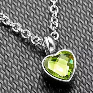 Op de afbeelding: Een zilveren ketting met een hartvormige hanger. De hanger heeft een lichtgroene edelsteen gezet in een zilveren rand. De ketting is een klassieke schakelstijl en de achtergrond is een gestructureerd, donkergrijs oppervlak.