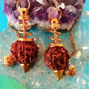 May include: Two gold-colored pendulums with a spiral design and a brown, textured, round weight. The pendulums are hanging from a gold chain and are resting on a purple amethyst crystal cluster.