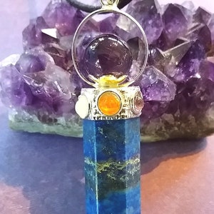 Puede incluir: Un colgante de cristal de lapisl&aacute;zuli azul con un engaste plateado y una esfera de cristal transparente que contiene un cristal de amatista p&uacute;rpura. El colgante est&aacute; rodeado de cristales m&aacute;s peque&ntilde;os de varios colores, incluyendo naranja, rojo y blanco.