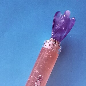May include: A decorative bottle stopper with a carved purple angel figure. The stopper has a silver band with a cut-out design and a pinkish-clear body with three small, round, pink embellishments. The background is a solid blue.