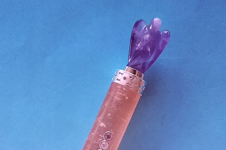 May include: A decorative bottle stopper with a carved purple angel figure. The stopper has a silver band with a cut-out design and a pinkish-clear body with three small, round, pink embellishments. The background is a solid blue.