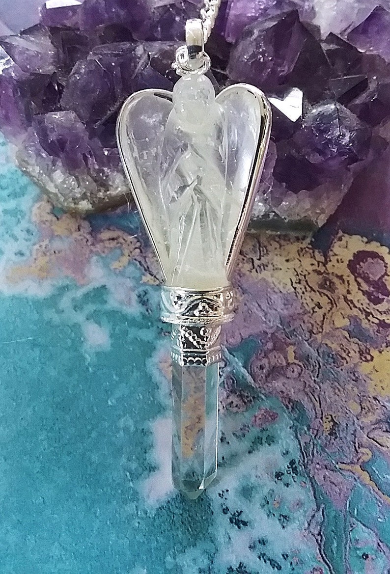 Op de afbeelding: Een heldere kwarts engelenvleugel hanger met een zilverkleurige metalen setting. De hanger hangt aan een zilveren ketting en rust op een bed van amethist kristallen.