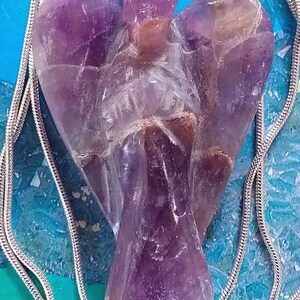 May include: A silver chain necklace with a purple amethyst angel pendant. The angel is carved from a single piece of amethyst and has a detailed design.