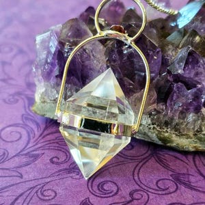 Puede incluir: Un colgante de cristal de cuarzo transparente de doble terminación, encerrado en un marco de metal plateado con una anilla circular en la parte superior. El colgante cuelga de una fina cadena plateada y presenta un diseño facetado y puntiagudo.