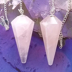 May include: Two pink rose quartz crystal pendulums with silver chains. The pendulums are faceted and have a pointed tip. They are hanging against a purple background.