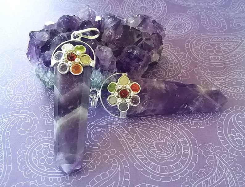 May include: Two amethyst crystal pendants with silver settings and colorful gemstone accents. The pendants are displayed on a bed of amethyst crystals.