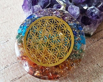 May include: A circular resin pendant featuring a gold-colored metal flower of life symbol embedded on the surface. The pendant contains layers of multi-colored crushed stones in red, orange, green, blue, and purple hues, with a silver-toned bail.