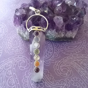 May include: A clear quartz crystal pendant with a silver setting and seven chakra stones. The stones are red, orange, yellow, green, blue, indigo, and violet. The pendant is resting on a bed of amethyst crystals.