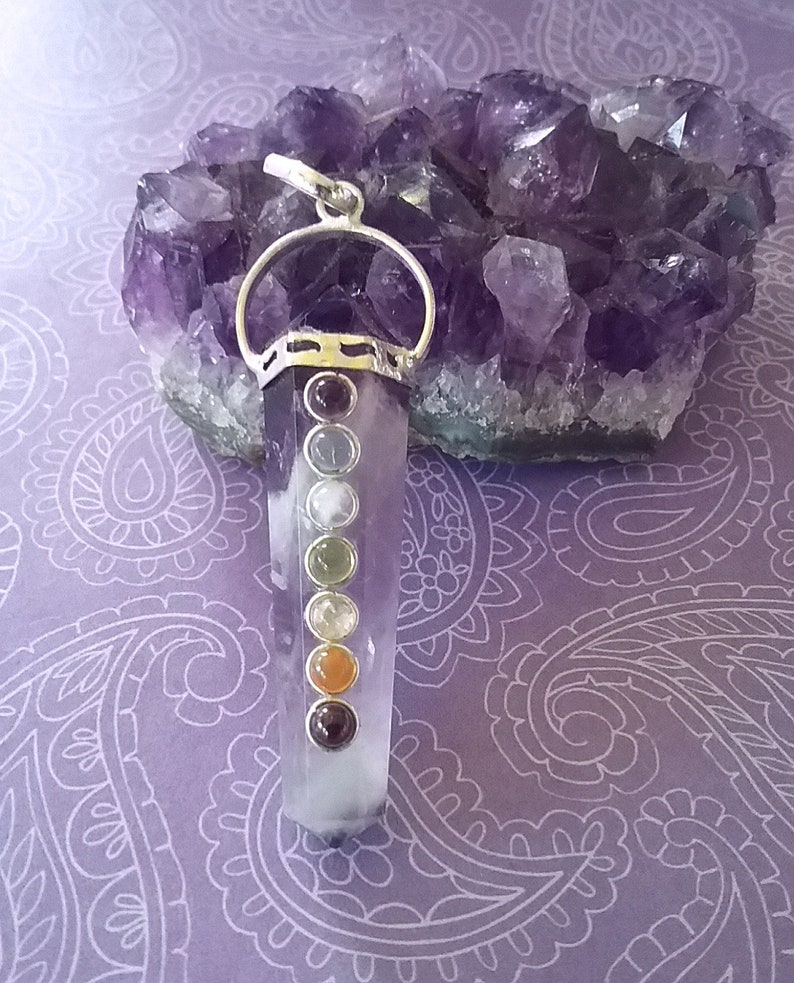 May include: A clear quartz crystal pendant with a silver setting and seven chakra stones. The stones are red, orange, yellow, green, blue, indigo, and violet. The pendant is resting on a bed of amethyst crystals.