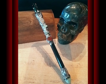 May include: A black wooden wand with silver accents and various coloured stones. The wand is decorated with a silver spiral design and has a green stone at the tip. The wand is lying on a wooden surface next to a black skull.